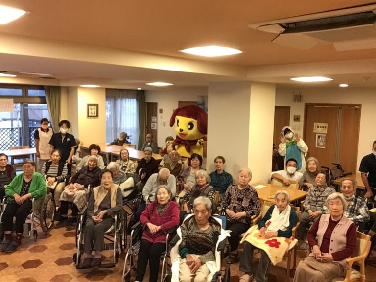 介護施設でのイベント行事。多数の入居者とキャラクターマスコット、スタッフが集まる共用スペース。